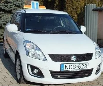 Fehér Suzuki Swift oktatóautó tetején a tanuló vezetőt jelző T táblával